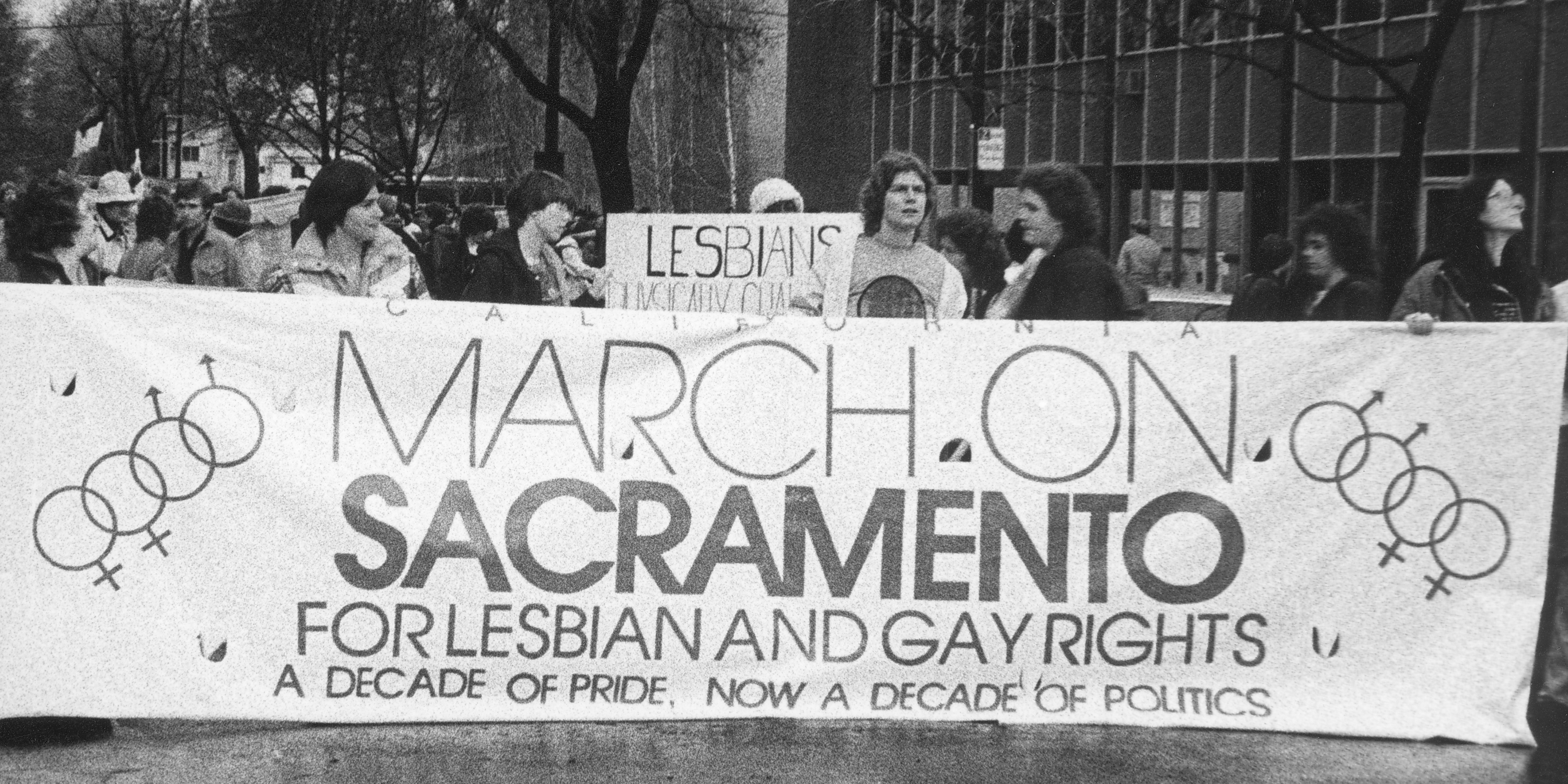 image from the 80s march in Sacramento for gay rights showing people holding a sign for the march on Sacramento.