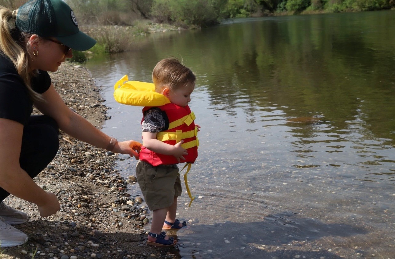 toddler in life jacket stepping into water with an adult behind him